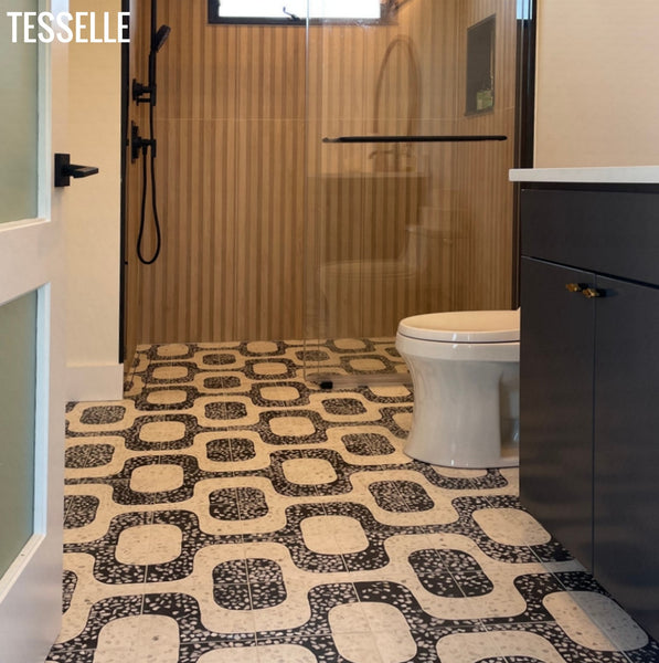 A bathroom interior showing a black and beige geometric patterned chunky terrazzo floor, a white toilet, a glass shower enclosure, and a dark gray vanity. The Tesselle brand name is visible in the top left corner.